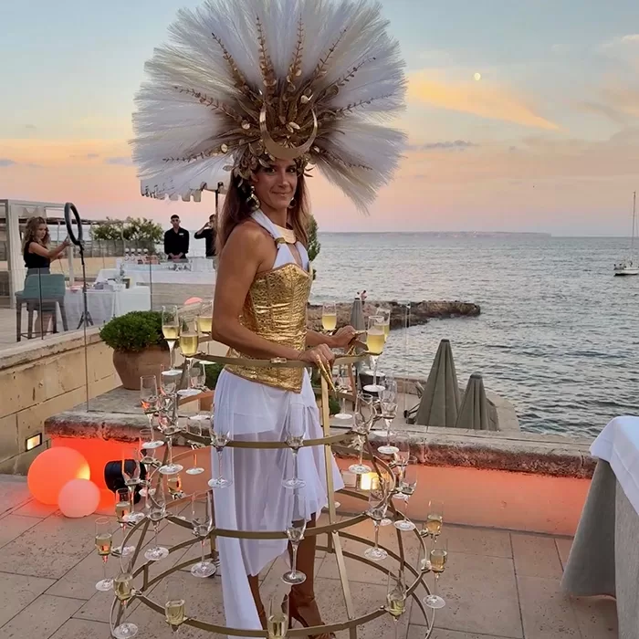 A woman in a gold and white costume with an elaborate headdress stands by the sea at sunset, serving champagne glasses from a circular metal frame attached to her waist. Guests mingle in the background on a terrace.