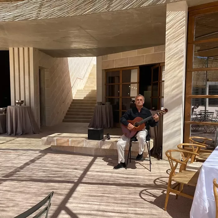 A man sits on a chair playing a guitar in a sunlit courtyard outside a building with large windows and stairs. Shadows from overhead create patterns on the ground and nearby tables and chairs.