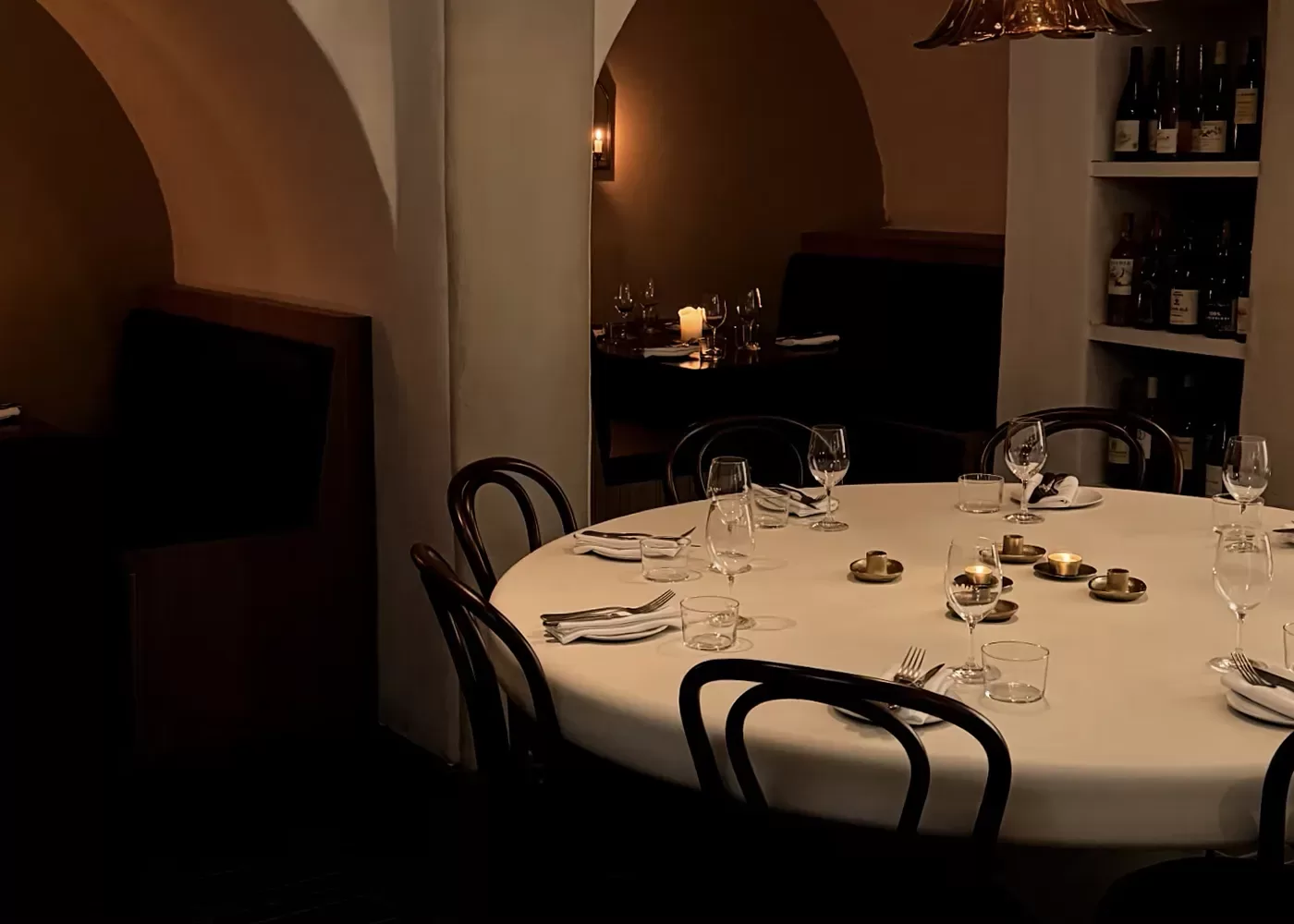 A dimly lit restaurant features a round table set with glasses, cutlery, napkins, and candles. There are dark wooden chairs around the table, and bottles of wine on shelves in the background. Booth seating is visible in an alcove.