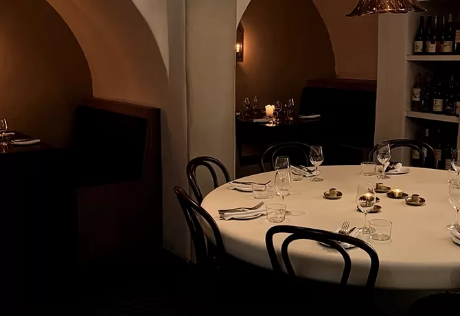 A dimly lit restaurant with a round table set for dining, featuring glassware, plates, and napkins. Built-in archway booths and shelves with wine bottles are visible in the background.