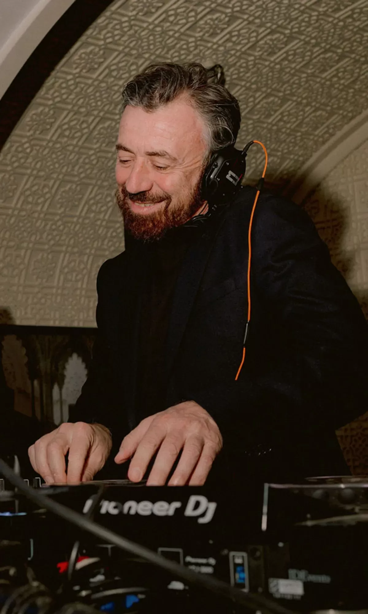 A man with a beard smiles while wearing headphones and using a Pioneer DJ controller in a warmly lit room with a patterned ceiling.