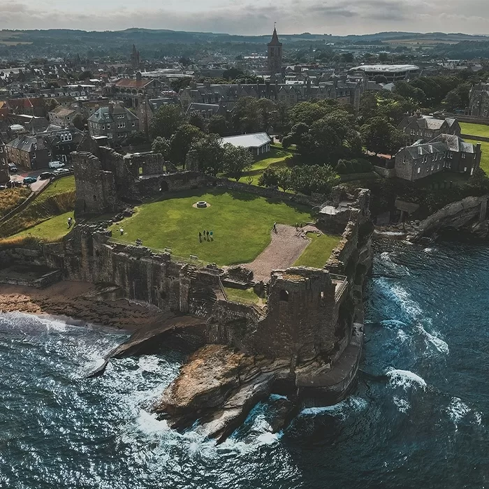 Aerial view of St Andrews Castle ruins perched on a rocky coastline, surrounded by green lawns and waves, with a town and distant hills in the background under a cloudy sky.