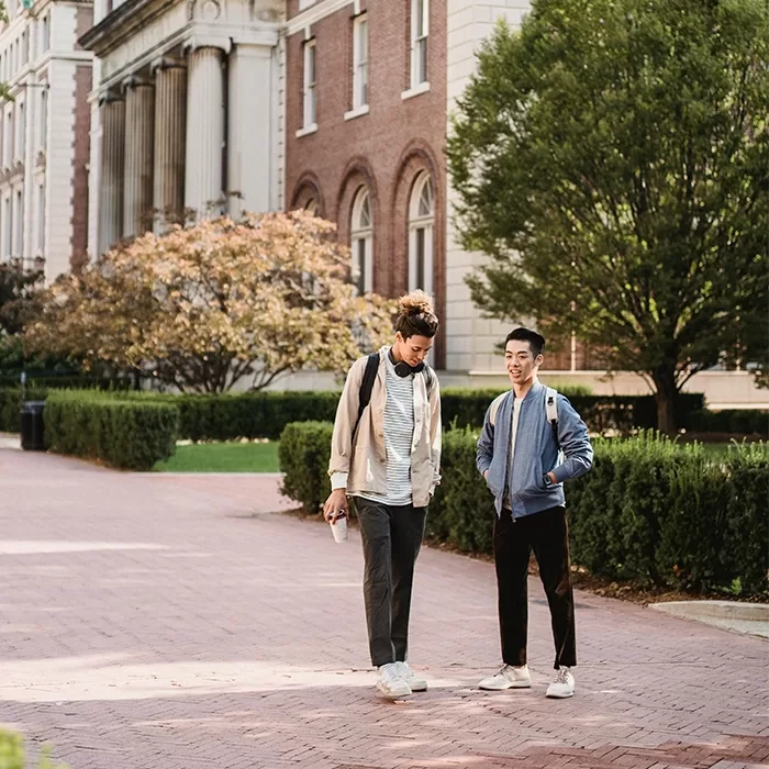 Two students with backpacks walk and talk on a brick path lined with trees and bushes, with a large academic building featuring columns and arched windows in the background.