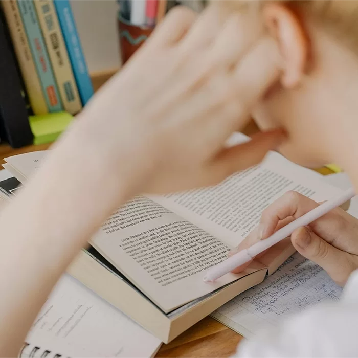 A person sits at a desk reading an open book, holding a pen and resting their head on one hand. Notes and other books are nearby, suggesting they are studying or doing homework.