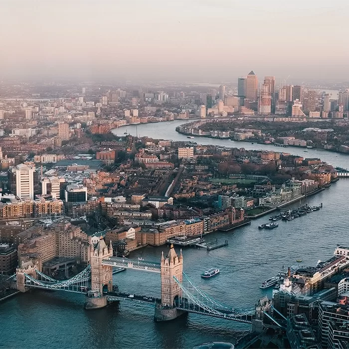 Aerial view of London at dusk, showing the Tower Bridge crossing the River Thames, with boats on the water and a backdrop of city buildings and skyscrapers under a hazy sky.