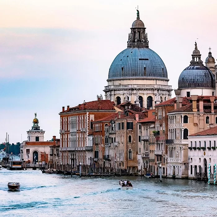 Venetian canal scene with boats on the water, historic buildings lining the canal, and the domed Basilica di Santa Maria della Salute in the background under a pastel sky.