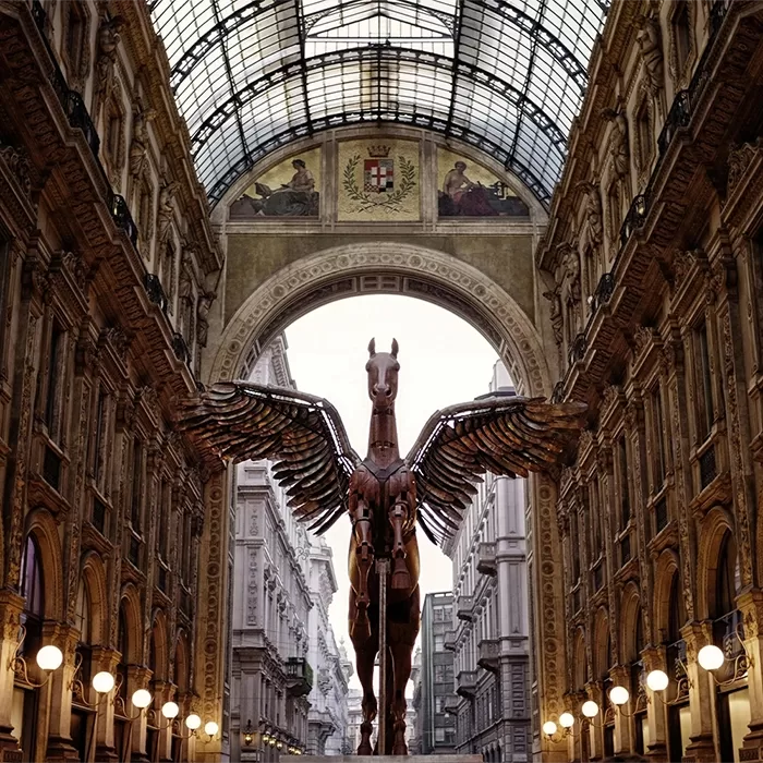 A large metallic Pegasus sculpture with outstretched wings stands beneath a glass-domed arcade, surrounded by ornate architecture and glowing lights in a grand indoor gallery.