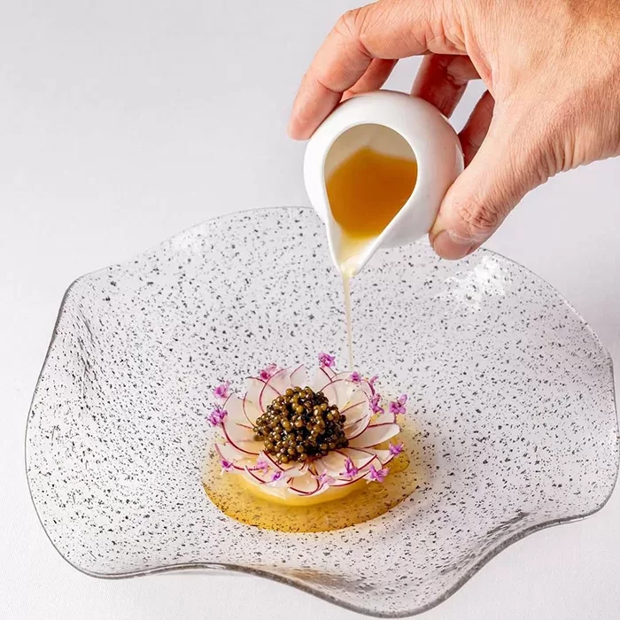 A hand pours sauce from a small white jug onto an elegant dish with caviar, thinly sliced radishes, edible flowers, and a circular yellow base, served on a speckled, wavy glass plate.