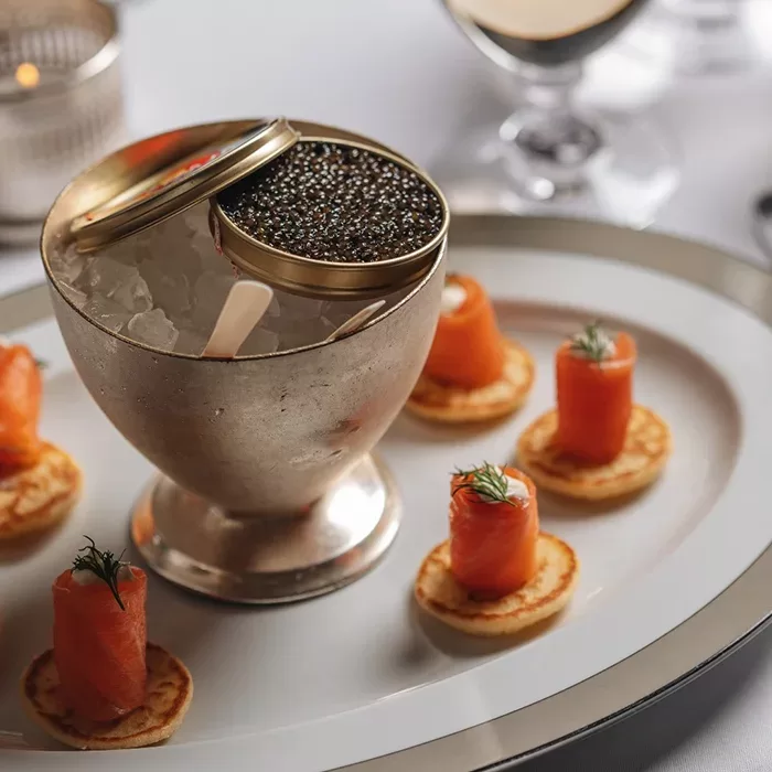 A tin of black caviar sits on ice in a silver dish, surrounded by blinis topped with rolled smoked salmon and dill, arranged neatly on a white plate.