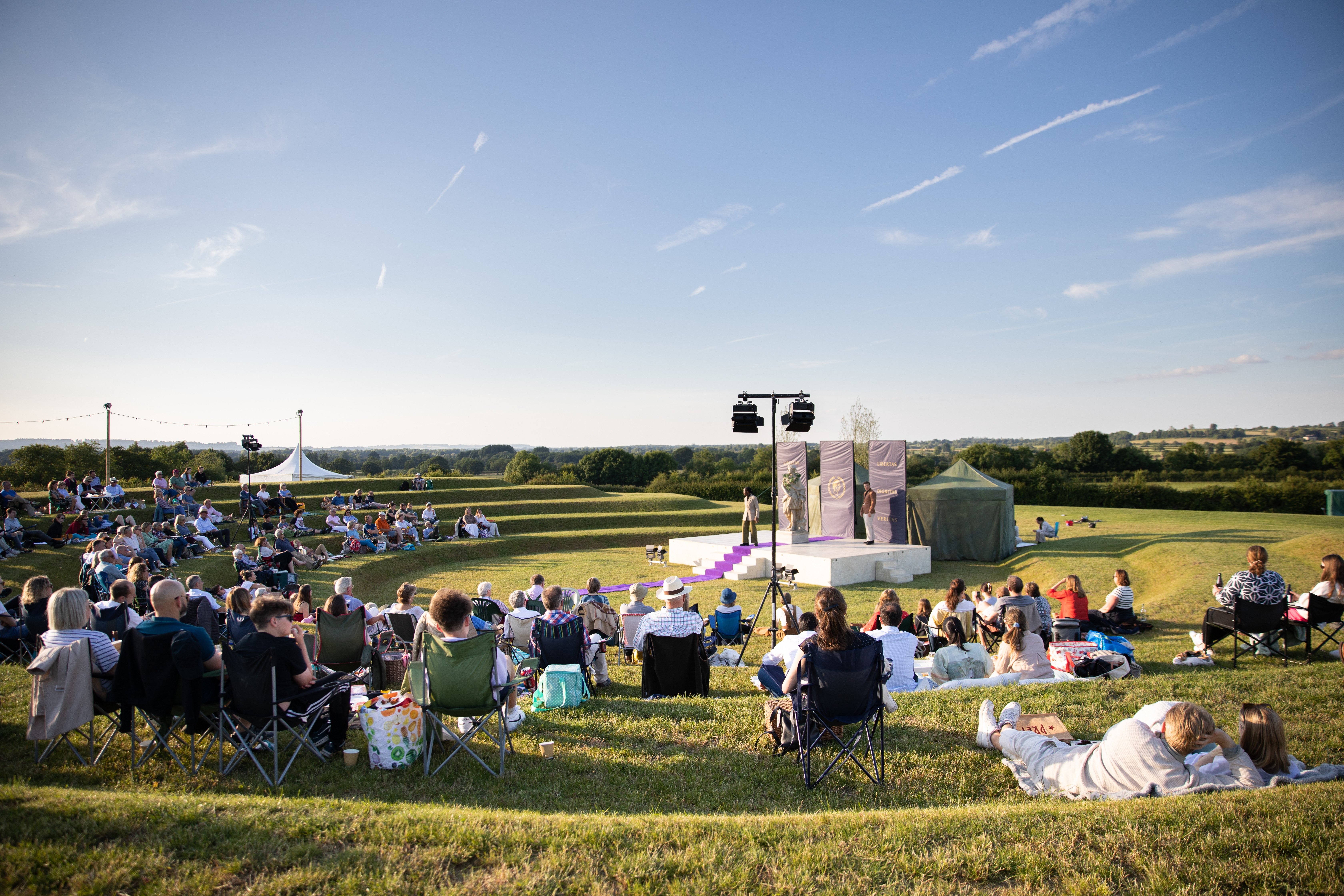 View above audience sat on rounded tiered grass watching white centre amphitheatre stage with man talking on top of purple carpet.