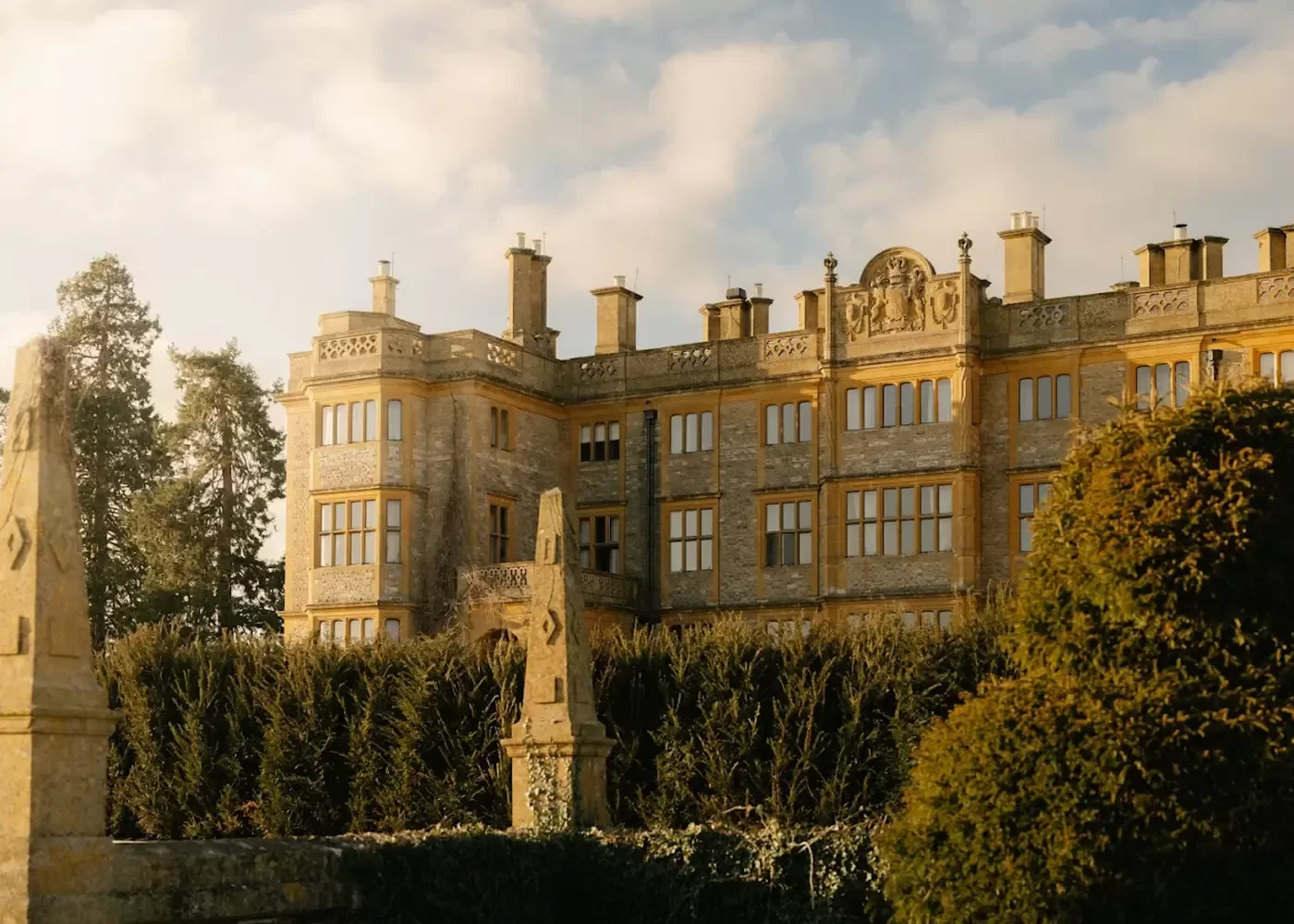 A grand stone mansion with many windows, ornate architectural details, and multiple chimneys stands behind tall hedges on a sunny day, with a lightly clouded sky above.