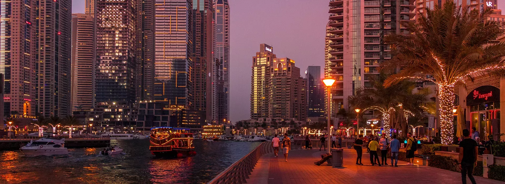 View of Dubai city at night time with busy wide streets, lit up tall buildings, palm trees and the sea filled with boats