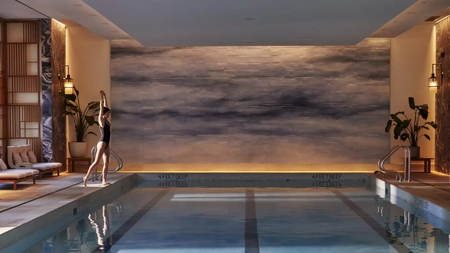 A person in a black swimsuit stands at the edge of an indoor swimming pool, stretching with arms raised. The pool area is softly lit, featuring a large, artistic wall mural and lounge chairs. Two potted plants are placed near the walls.