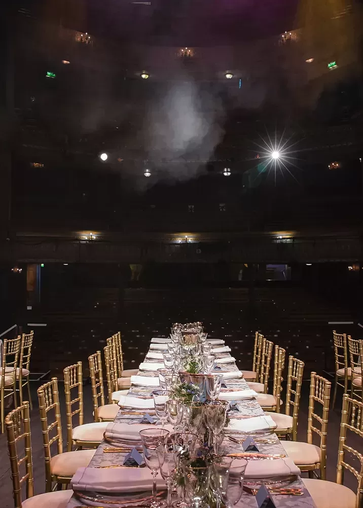 A long, elegant dining table is set up on a theatre stage, adorned with glassware, silverware, and floral centrepieces. The table is surrounded by gold chairs. Theatre seats and dim lighting are visible in the background, with a spotlight shining onto the stage.