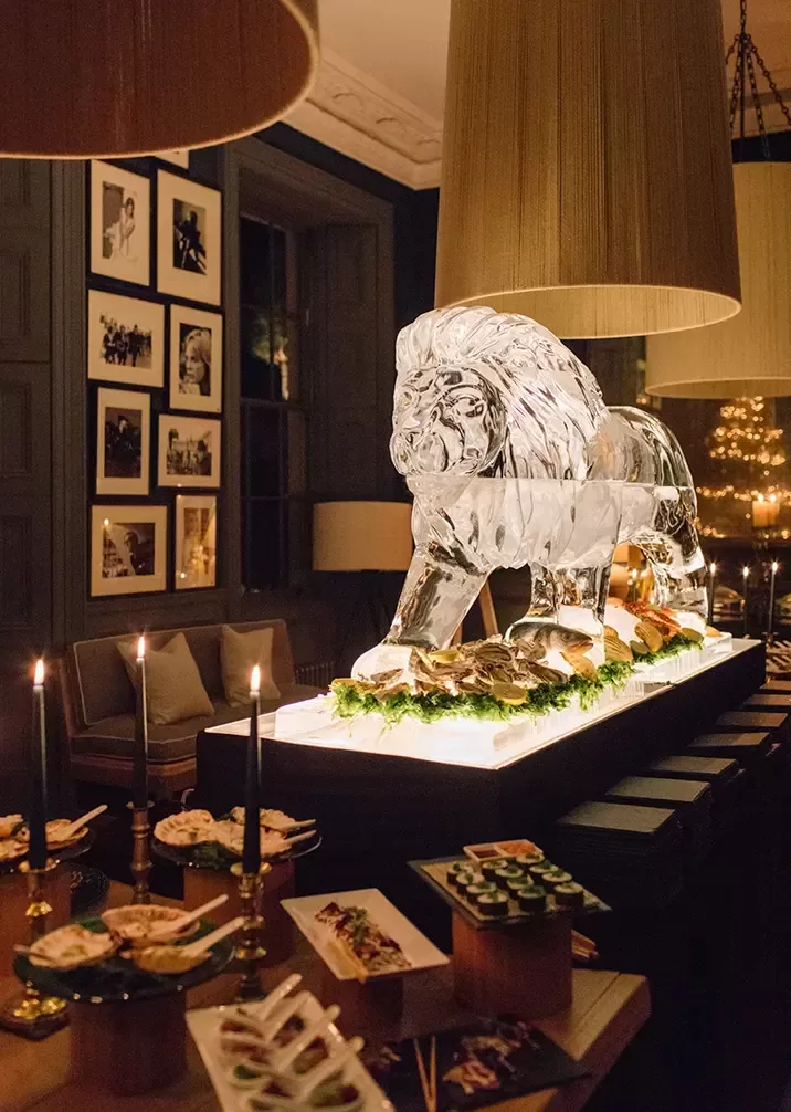 A grand ice sculpture of a lion dominates the centre of a dimly lit room, with various delicacies and greens displayed around it. Several lit candles and framed photos on the wall provide a touch of warmth and elegance to the sophisticated setting.