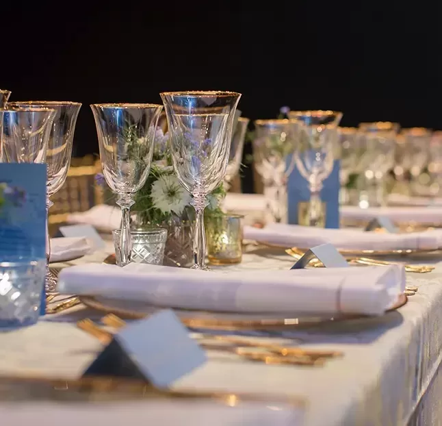 A close-up of an elegantly set dining table with folded white napkins, gold-rimmed glasses, and luxurious tableware. Small floral arrangements and candles in glass holders add to the sophisticated ambiance. Place cards are positioned at each setting.