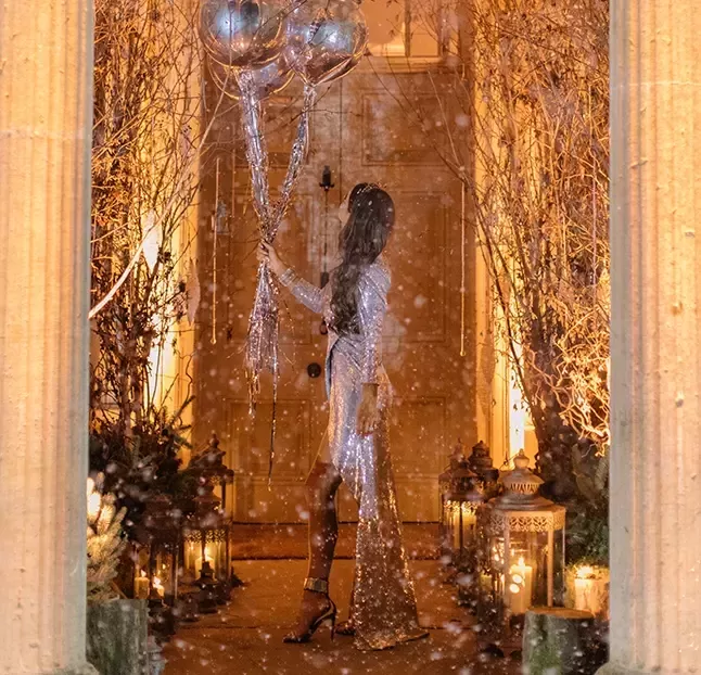 A person in a silver outfit stands on a festive, illuminated path holding silver balloons. The path is lined with decorative lanterns and trees adorned with lights. Snow is lightly falling, creating a magical winter scene.