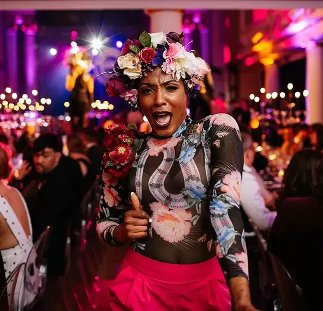 A woman wearing a vibrant floral headpiece, smiling joyfully at a lively party atmosphere.