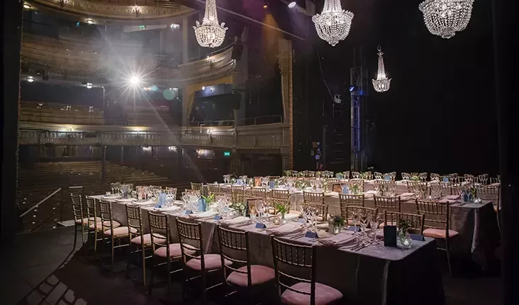 A grand theatre with ornate chandeliers and balconies is set up for a formal dinner event. The stage is arranged with long banquet tables elegantly decorated with white tablecloths, floral centerpieces, glassware, and silverware, creating a sophisticated dining atmosphere.