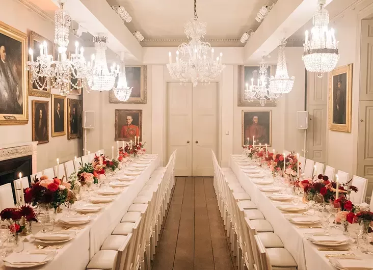 A grand dining hall with a long, elegantly set table adorned with red and white floral arrangements and candles. Portrait paintings hang on the walls, and multiple glittering chandeliers illuminate the room, creating a luxurious atmosphere.