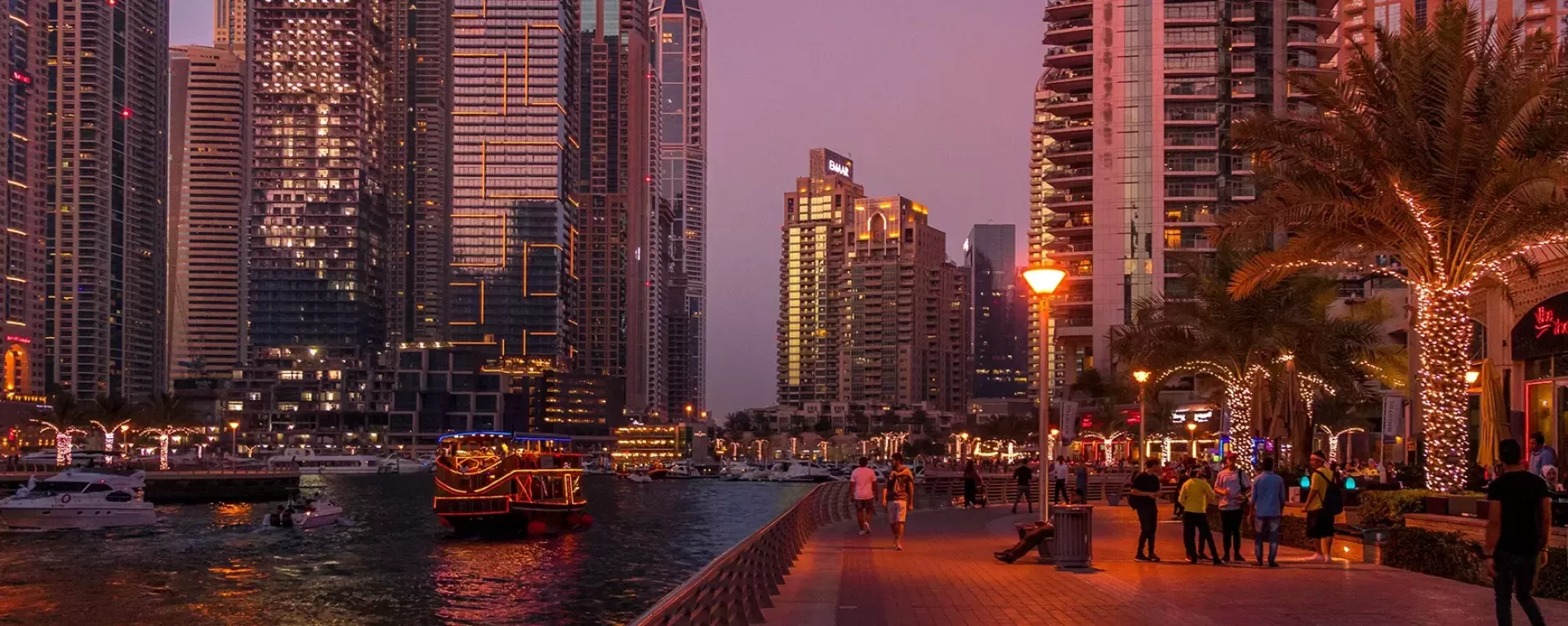 View of Dubai city at night time with busy wide streets, lit up tall buildings, palm trees and the sea filled with boats