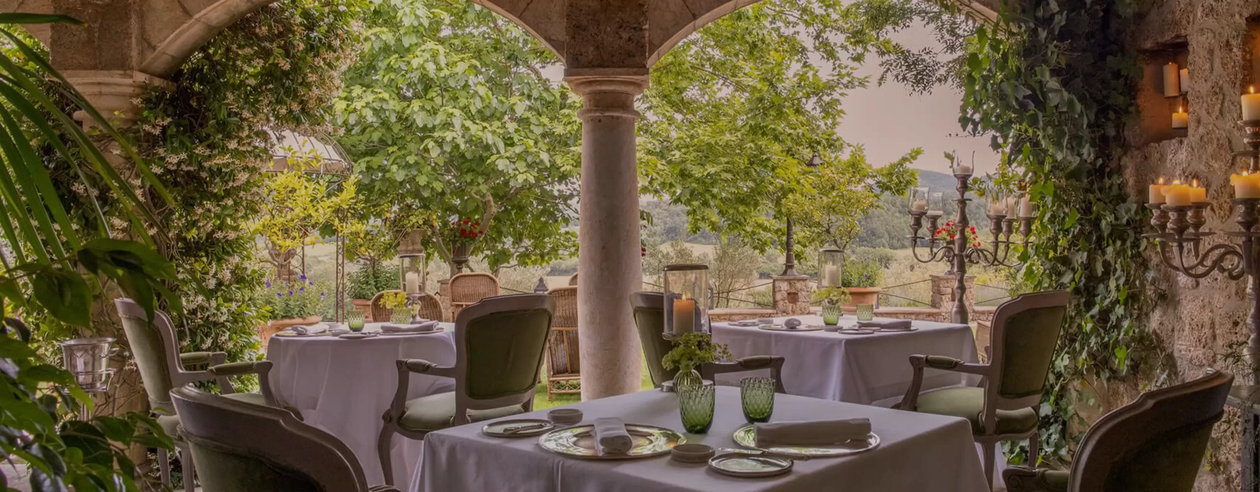Elegant outdoor restaurant patio with white-tableclothed tables, green cushioned chairs, greenery-covered stone arches, and lush garden views. Candleholders and glassware add a romantic, serene atmosphere.
