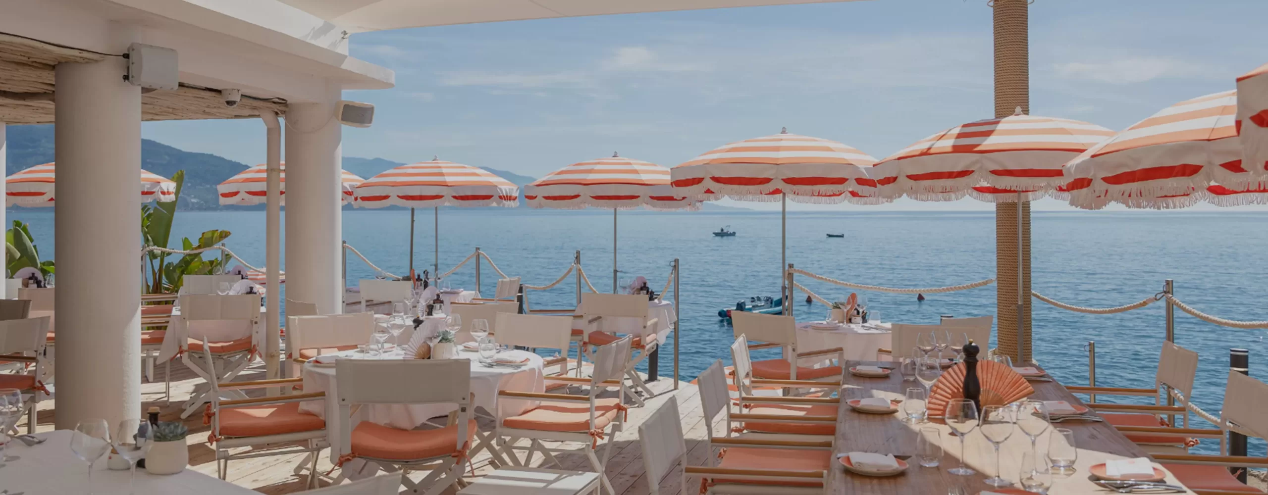 Tables and chairs set for dining on a seaside terrace with orange and white striped umbrellas, overlooking a calm blue sea and distant boats under a sunny, clear sky.