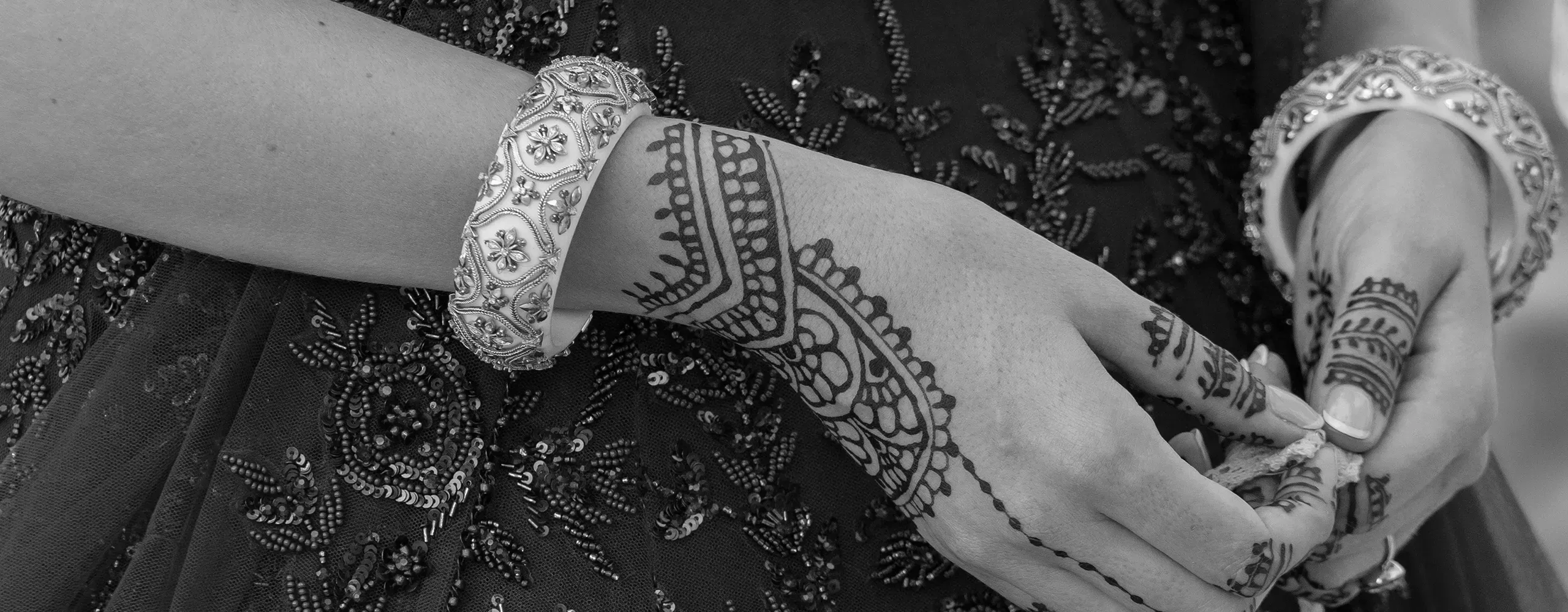 A close-up of hands decorated with intricate henna designs, wearing ornate bangles, against a dark, embroidered fabric background.