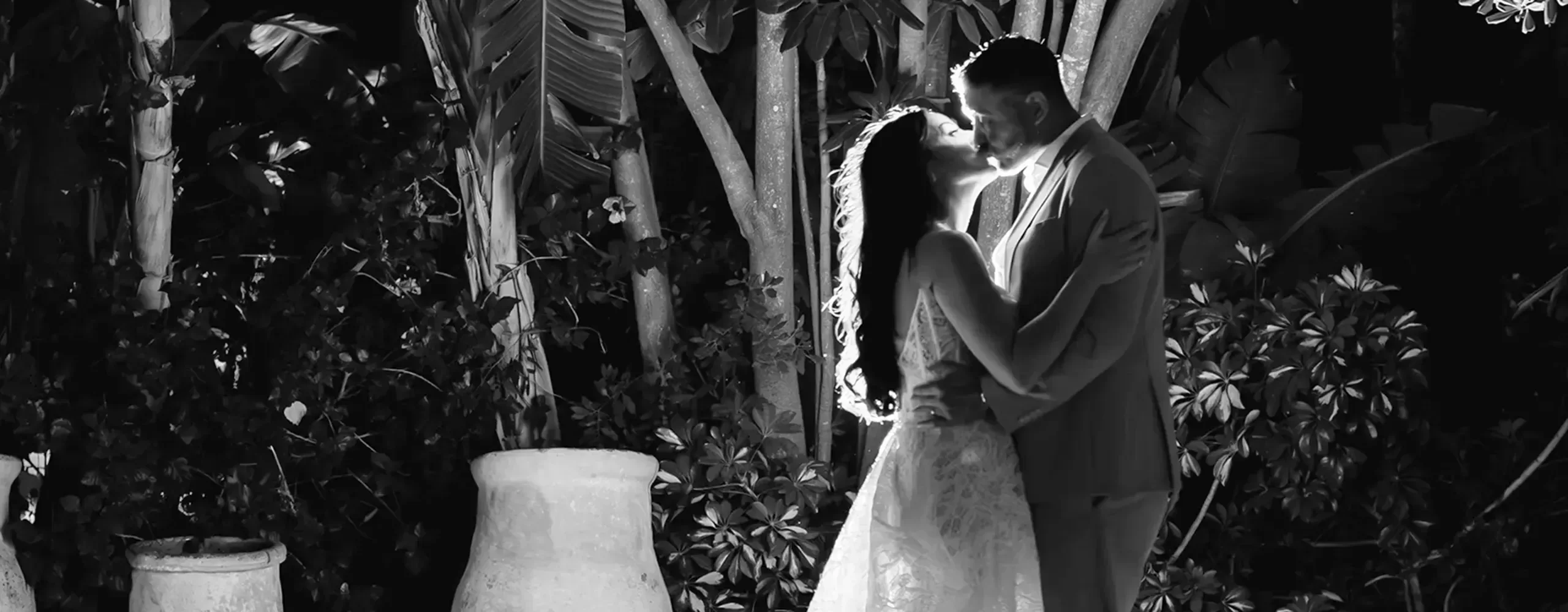 A bride and groom share a romantic kiss at night, surrounded by lush plants and trees, with soft lighting highlighting their embrace.