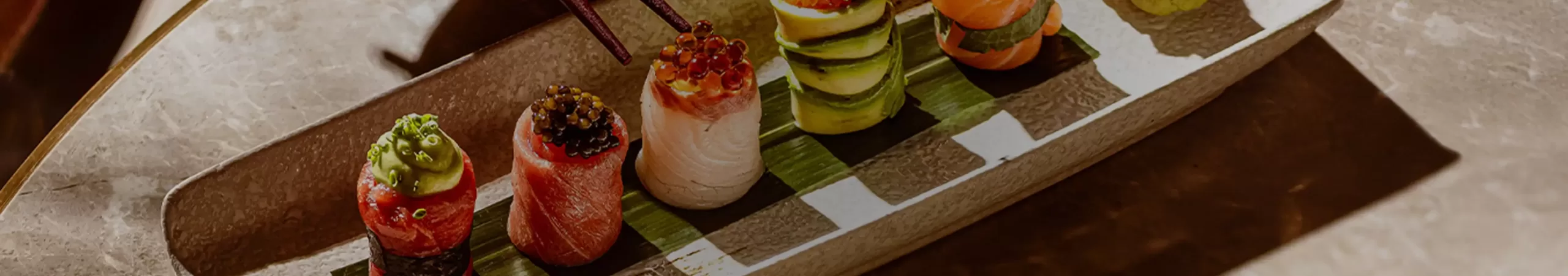 A rectangular plate with six pieces of colorful sushi, each topped with various ingredients, garnished with herbs. A hand holds chopsticks, picking up one sushi piece. Wasabi and pickled ginger are at the end of the plate.