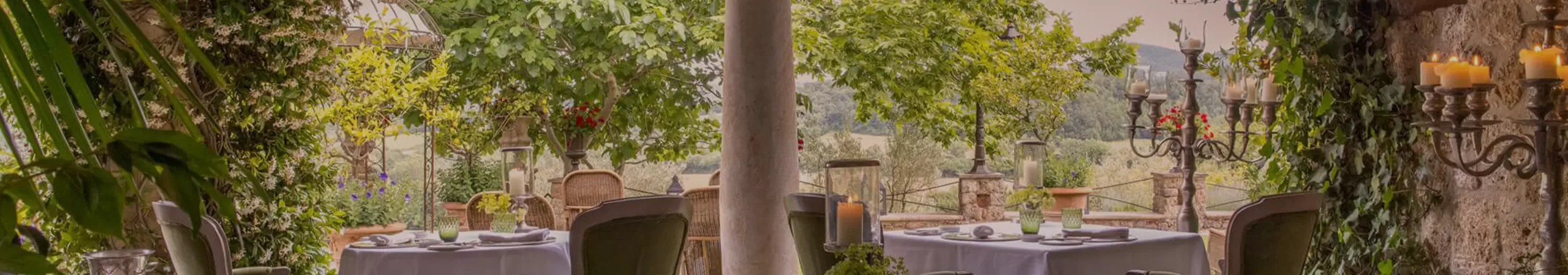 Elegant outdoor restaurant patio with white-tableclothed tables, green cushioned chairs, greenery-covered stone arches, and lush garden views. Candleholders and glassware add a romantic, serene atmosphere.