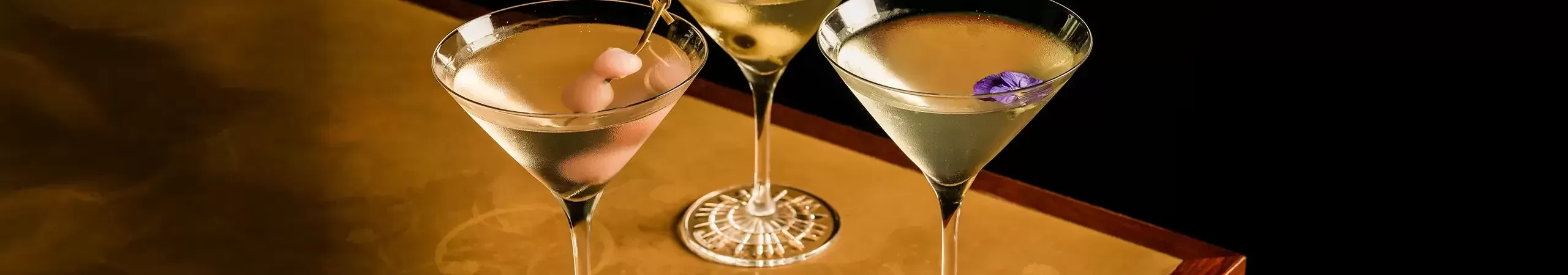 Three martini glasses on a gold table, each with a different garnish: one with olives, one with a cocktail onion, and one with a purple flower. Liquid is being poured into the glass with olives.