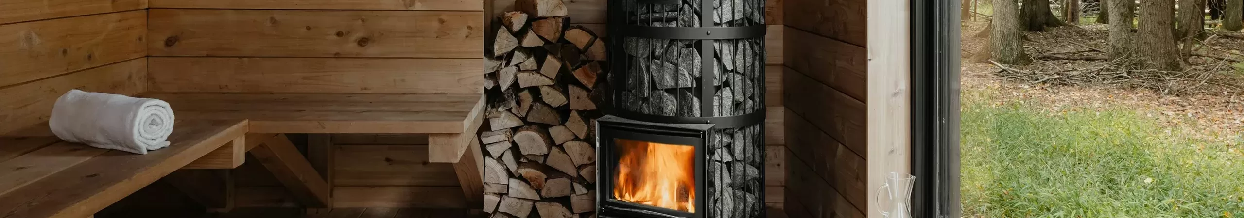 A cosy wooden sauna interior with a burning wood stove, stacked firewood, a rolled towel on a bench, a bucket with a ladle, and a large window showing a forest outside.