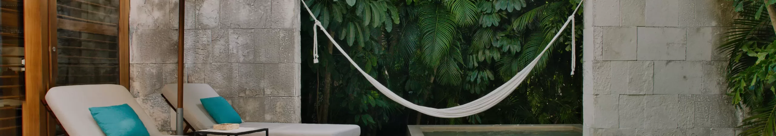 Two cushioned lounge chairs with teal pillows sit under a large umbrella beside a small pool. A white hammock hangs over the water, and lush green tropical plants cover the background wall.