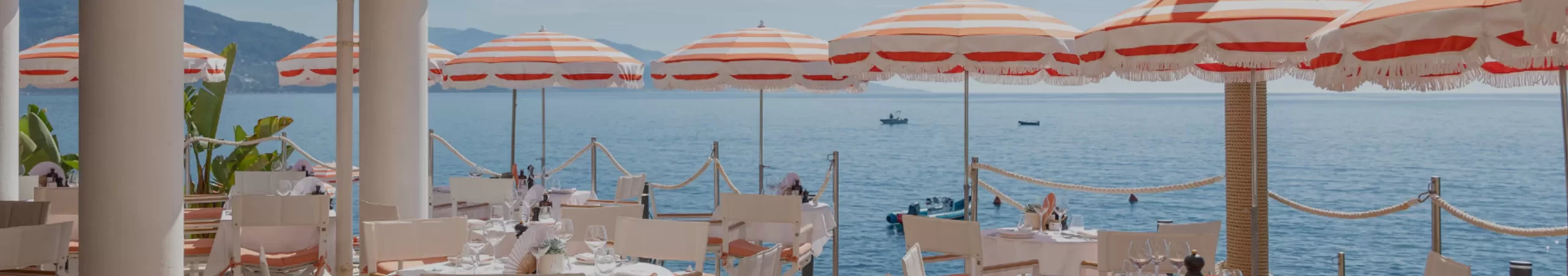 Tables and chairs set for dining on a seaside terrace with orange and white striped umbrellas, overlooking a calm blue sea and distant boats under a sunny, clear sky.