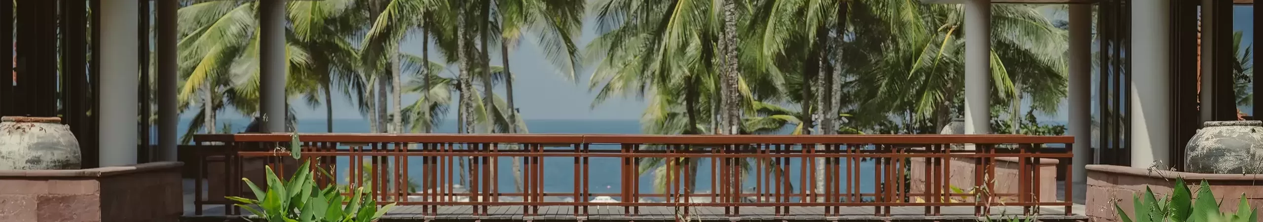A wooden bridge spans over a pond, surrounded by lush tropical plants and palm trees, with a view of the ocean in the background between two buildings.