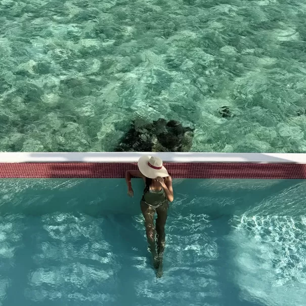 A woman in a sunhat and swimsuit stands in a swimming pool, leaning against the edge with clear blue ocean water visible beyond the pool.