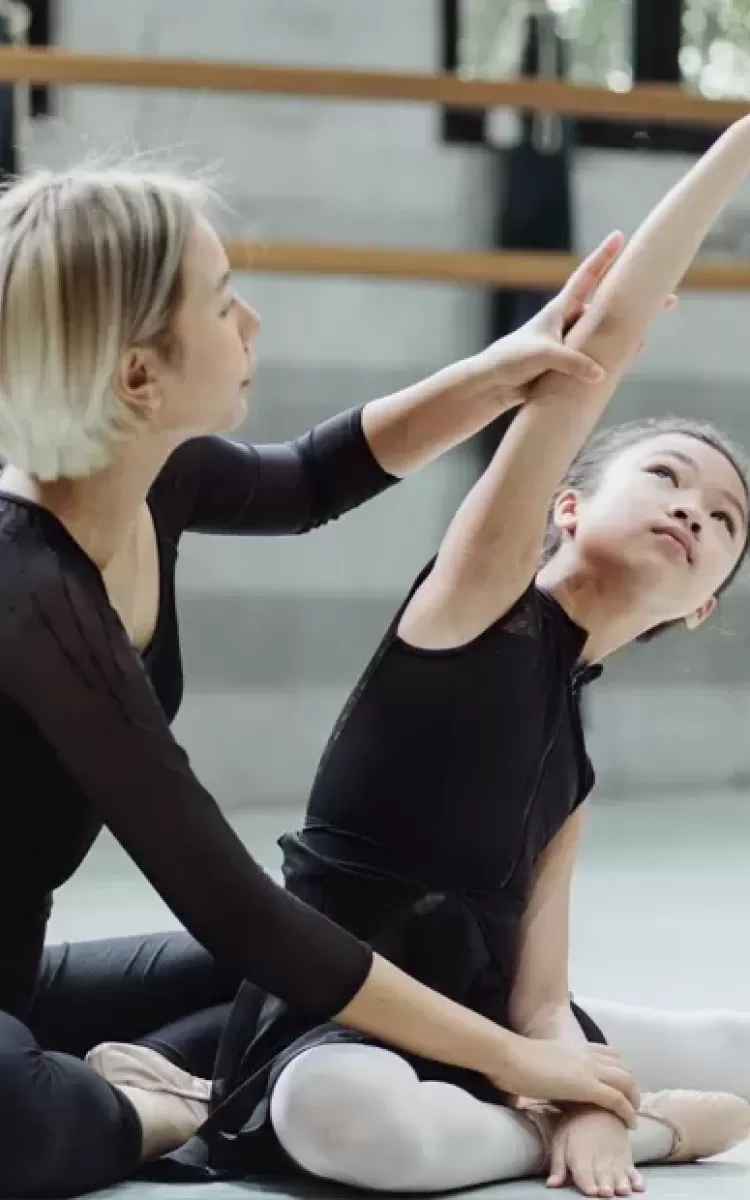 Ballet teacher sat on floor with child student helping her with a pose