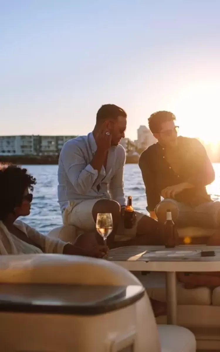 Group of people sat on the back of a luxury yacht at sea drinking wine and talking with sunset in the background
