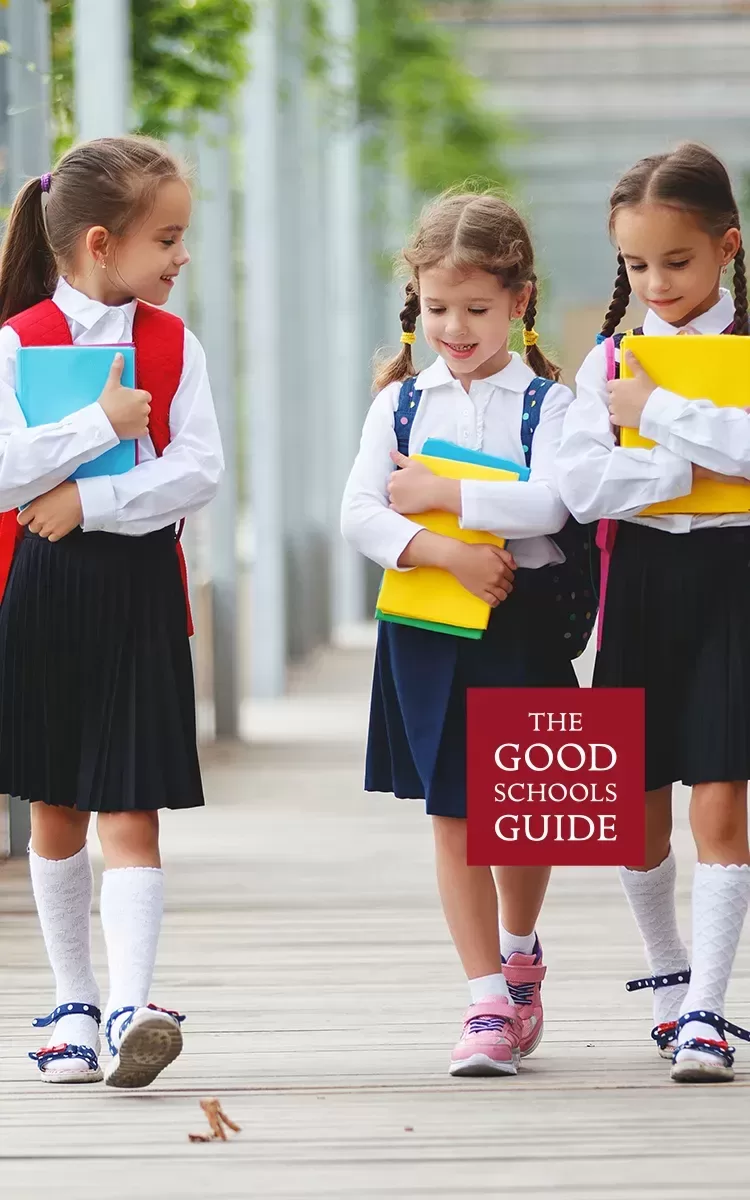 Three young girls in school uniforms walk together outdoors, smiling and holding colorful books. They have backpacks and are engaged in conversation. The text The Good Schools Guide appears in the lower right corner.