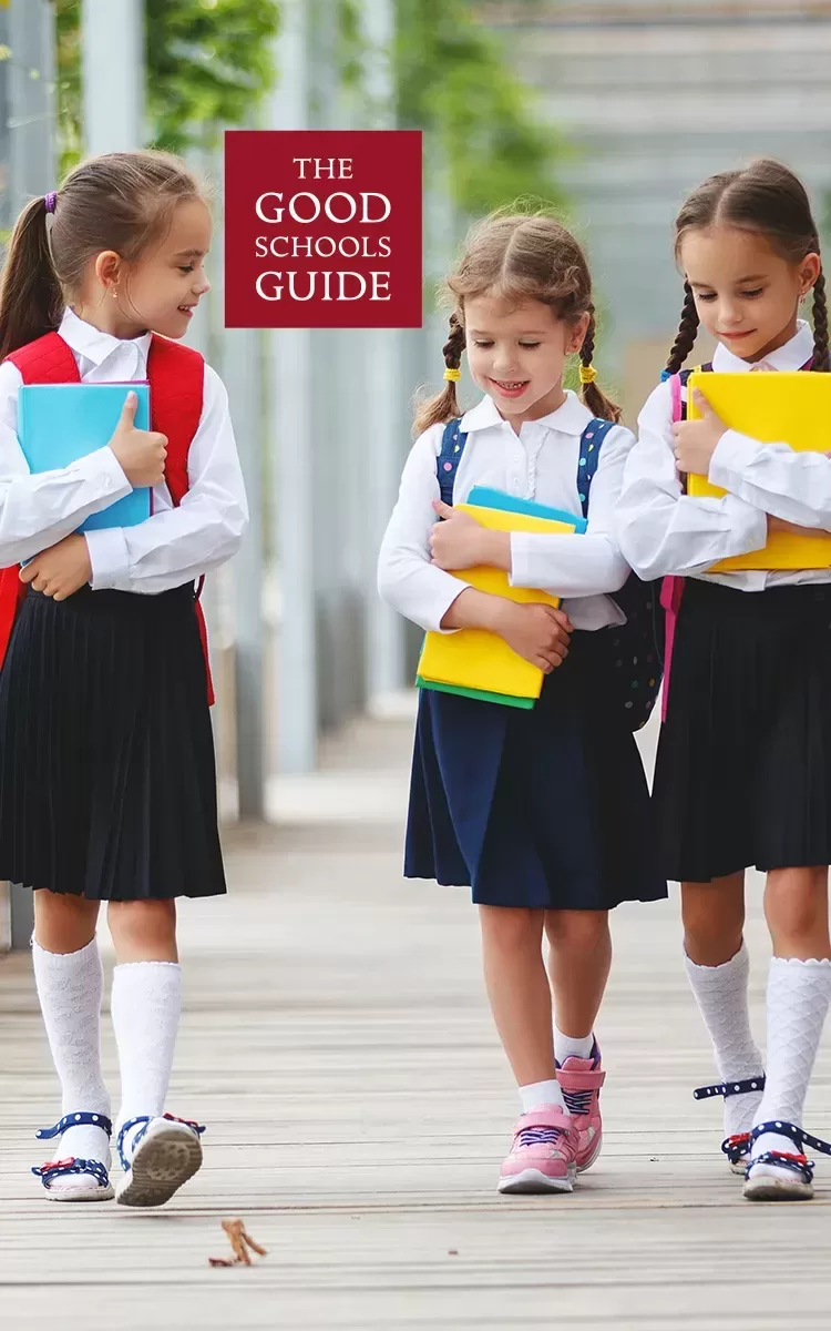 Three young girls in school uniforms walk outdoors, smiling and carrying colorful books. The text THE GOOD SCHOOLS GUIDE appears above them.