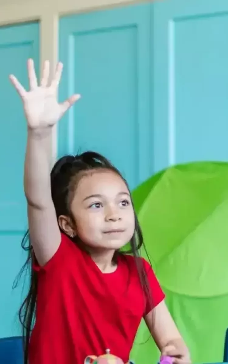 Four young children in a classroom sit at a table, three with hands raised, smiling and looking toward a teacher standing in front of them. Blue cabinets and a green board are visible in the background.