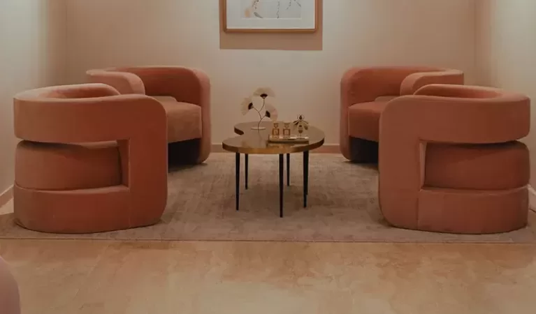 A modern lounge with peach-colored armchairs, a small coffee table with decor items, soft lighting, light walls and floor, and abstract artwork on the back wall.