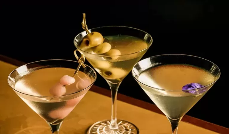 Three martini glasses on a table, each with a different garnish: cocktail onions, green olives, and a purple flower. The background is dark, highlighting the drinks.