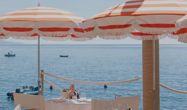 Outdoor dining area by the sea with tables set for a meal, featuring orange-and-white striped umbrellas, wooden chairs, and a view of boats and people swimming in the blue water under a clear sky.