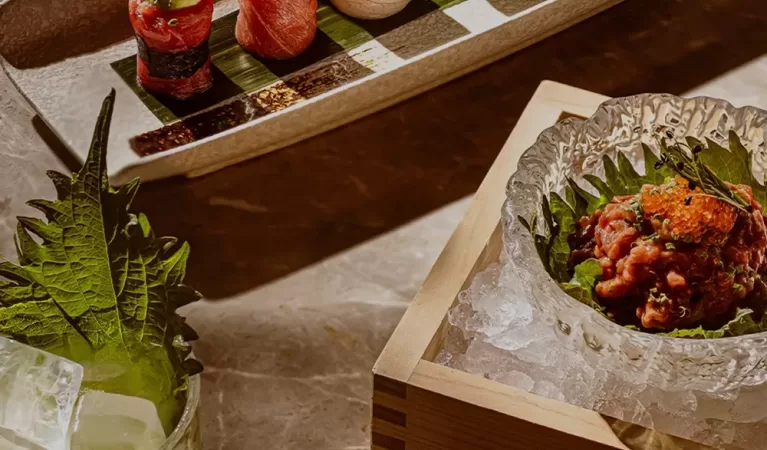 A hand holds chopsticks over a platter of colorful sushi rolls, with a cocktail, a bowl of seafood on ice, and a dish of fried food on a marble table in warm lighting.