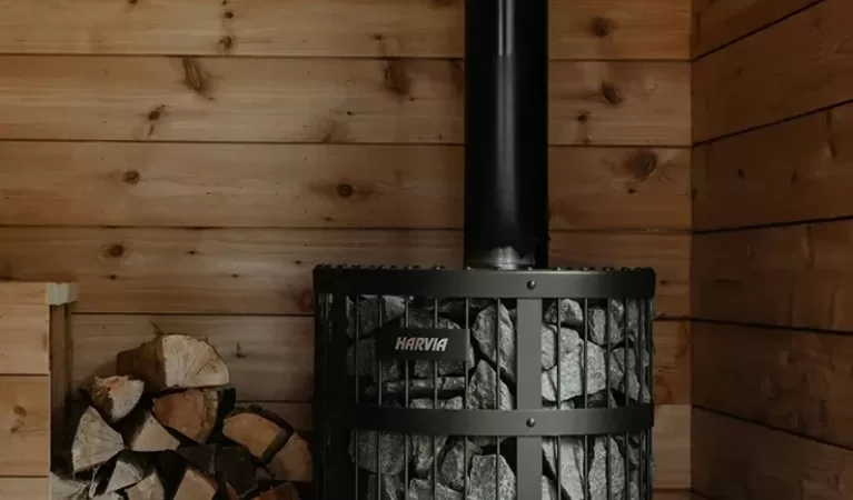 A wood-burning stove with a metal chimney stands in a wooden room, surrounded by stacked firewood and a wooden bucket with a ladle on the floor nearby. The stove emits a bright, warm fire.