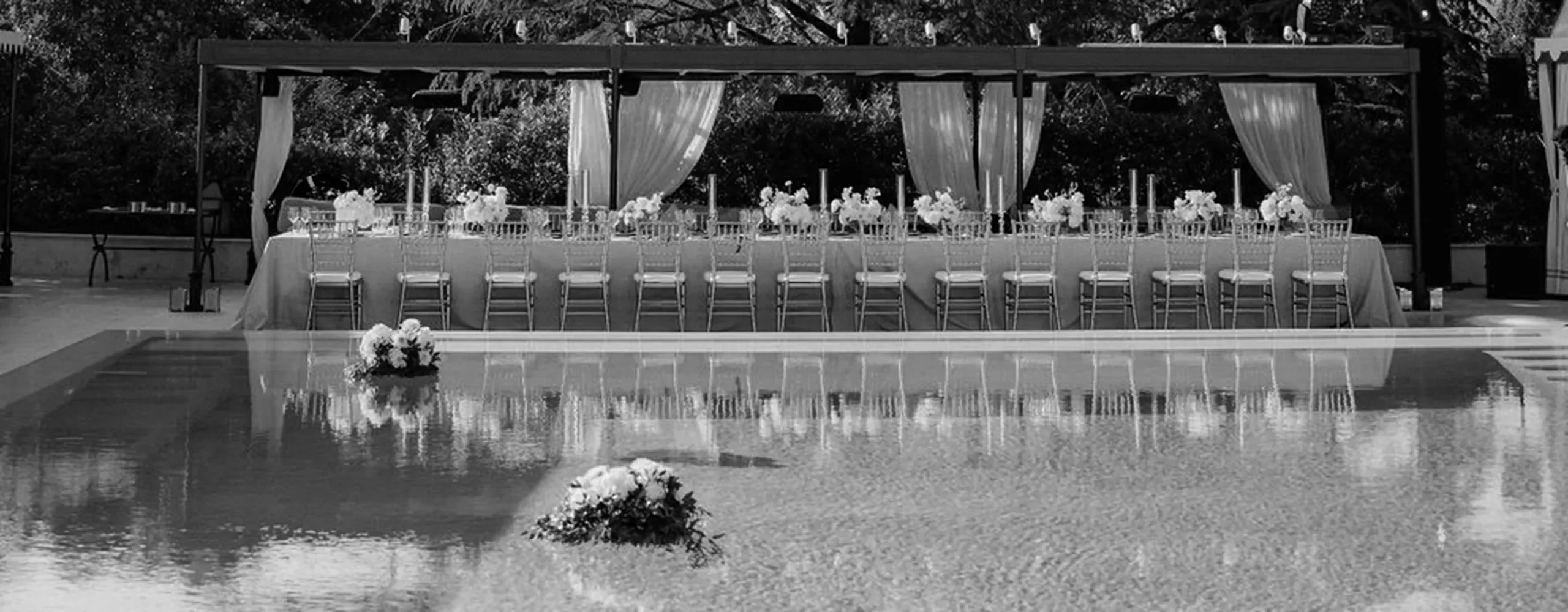 Long banquet table with elegant chairs and flower arrangements set beside a reflective pool, under a canopy with draped curtains; outdoor setting with trees in the background. Black and white image.