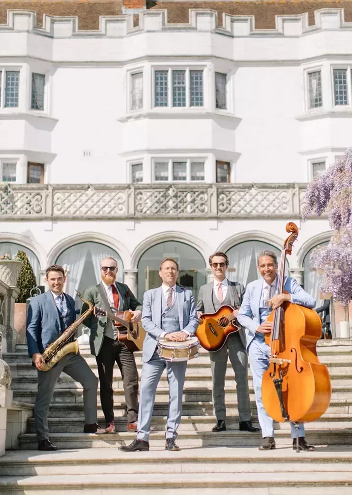 A band poses for a photo on the grand steps of a mansion, showcasing their stylish outfits and camaraderie.