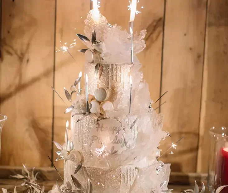 A three-tiered, elegantly decorated birthday cake adorned with sparklers and white foliage. The cake features a snow-like design with intricate icing details and subtle metallic accents against a rustic wooden background, creating a whimsical, celebratory ambiance.
